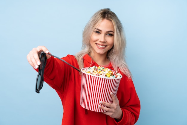 Chica adolescente sobre azul aislado con gafas 3d y sosteniendo un gran cubo de palomitas de maíz