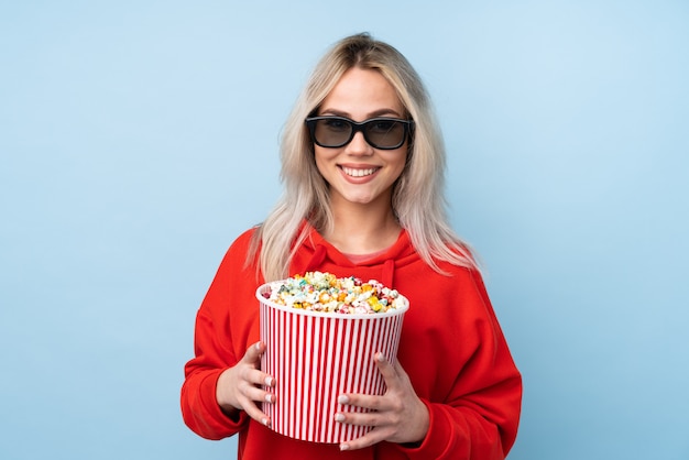 Chica adolescente sobre azul con gafas 3d y sosteniendo un gran cubo de palomitas de maíz