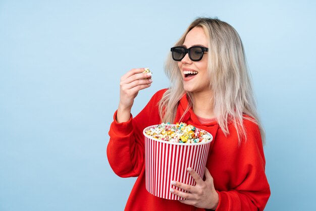 Chica adolescente sobre fondo azul aislado con gafas 3d y sosteniendo un gran cubo de palomitas de maíz