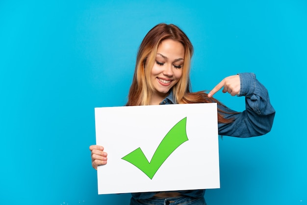 Chica adolescente sobre fondo azul aislado sosteniendo un cartel con el icono de marca de verificación verde de texto y apuntando