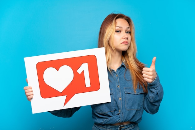 Foto chica adolescente sobre fondo azul aislado sosteniendo un cartel con el icono me gusta con el pulgar hacia arriba