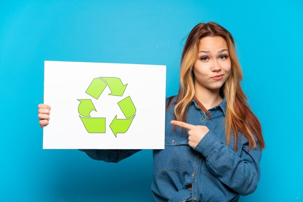 Chica adolescente sobre fondo azul aislado sosteniendo un cartel con el icono de reciclaje y apuntándolo