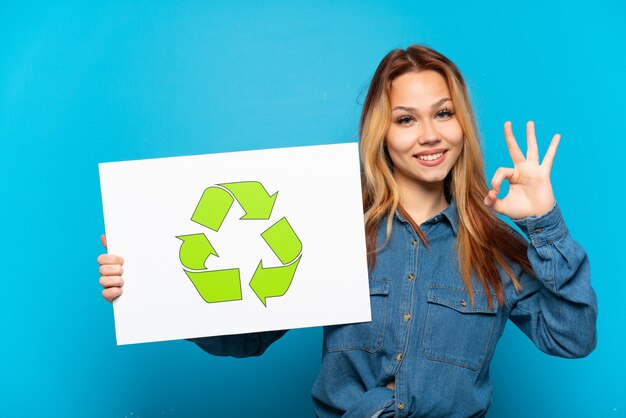 Chica adolescente sobre fondo azul aislado sosteniendo un cartel con el icono de reciclaje y celebrando una victoria