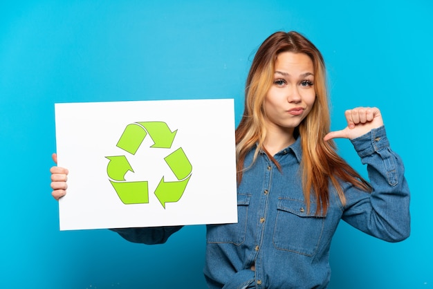 Chica adolescente sobre fondo azul aislado sosteniendo un cartel con el icono de reciclaje con gesto orgulloso