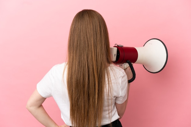 Chica adolescente sobre fondo rosa aislado sosteniendo un megáfono y en posición trasera