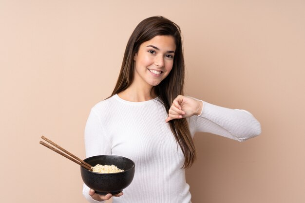 Chica adolescente sobre pared aislada orgullosa y satisfecha mientras sostiene un tazón de fideos con palillos