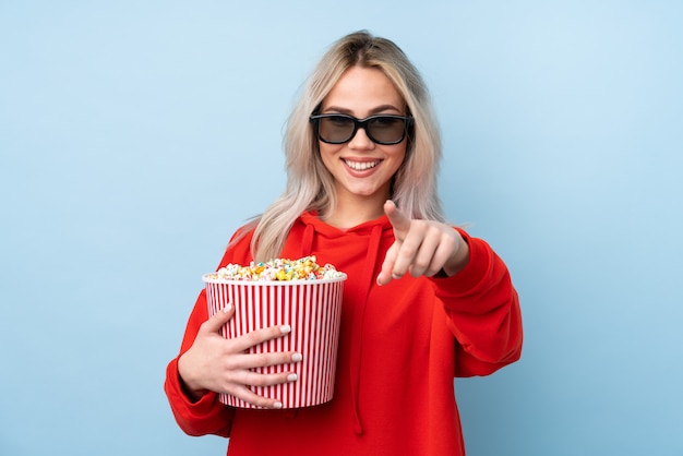 Chica adolescente sobre pared azul aislada con gafas 3d y sosteniendo un gran cubo de palomitas de maíz mientras apunta hacia adelante