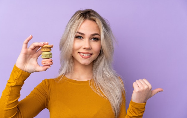 Foto chica adolescente sobre pared púrpura con coloridos macarons franceses y apuntando hacia el lado