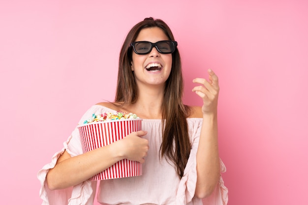 Chica adolescente sobre pared rosa aislada con gafas 3d y sosteniendo un gran cubo de palomitas de maíz mientras apunta hacia adelante