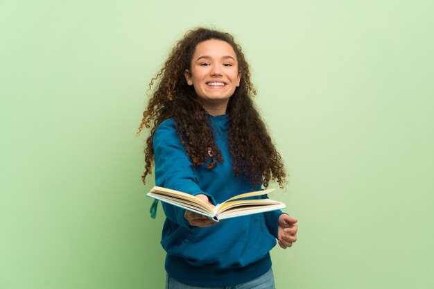 Chica adolescente sobre pared verde sosteniendo un libro y dárselo a alguien