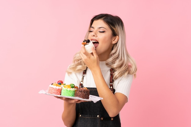 Chica adolescente sobre rosa aislado con mini tortas y comerlo