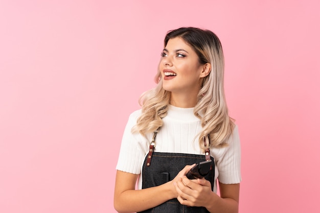 Chica adolescente sobre rosa aislado con peluquería o vestido de barbero y sosteniendo la cortadora de cabello