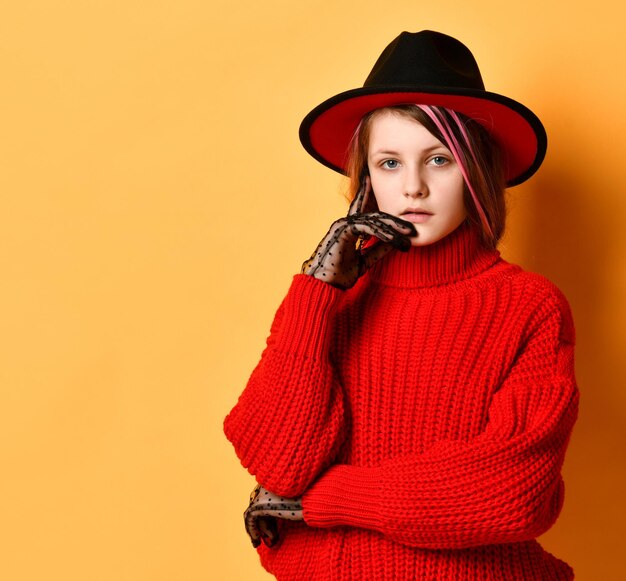 Chica adolescente con un sombrero fedora y un suéter rojo brillante que está sobre un fondo de estudio amarillo