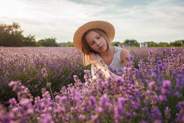 Chica adolescente soñadora con sombrero de paja se sienta en la belleza del campo de lavanda de la naturaleza, estilo de vida de verano