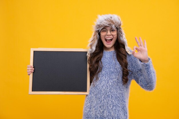 Chica adolescente sostenga en un cálido sombrero de invierno sostenga pizarra pizarra con espacio de copia sobre fondo amarillo Cara emocionada emociones alegres de chica adolescente