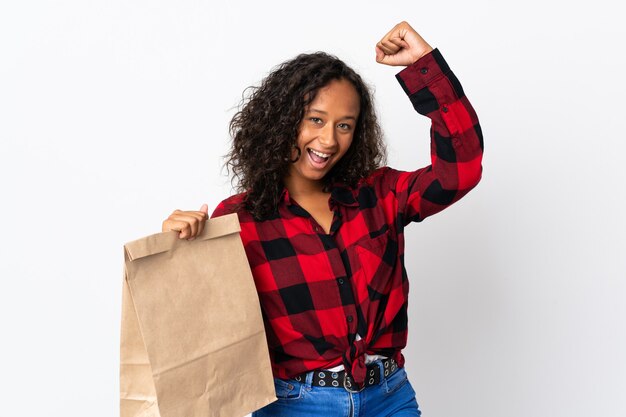 Chica adolescente sosteniendo una bolsa de compras para llevar aislado en la pared blanca celebrando una victoria