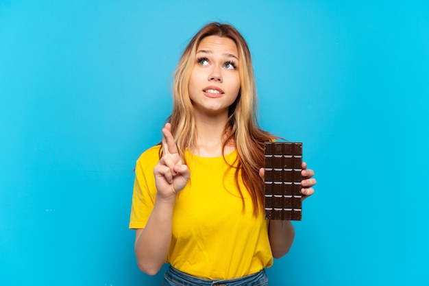 Chica adolescente sosteniendo chocolat sobre fondo azul aislado con los dedos cruzando y deseando lo mejor