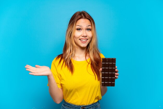 Chica adolescente sosteniendo chocolat sobre fondo azul aislado con expresión facial conmocionada