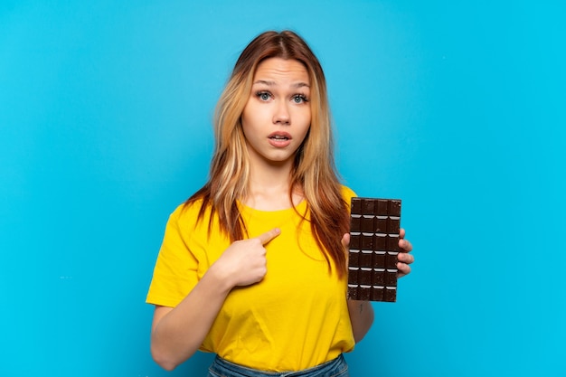 Chica adolescente sosteniendo chocolat sobre fondo azul aislado con expresión facial sorpresa