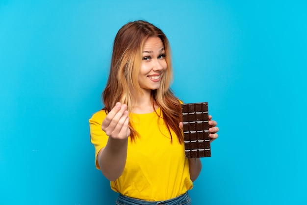 Chica adolescente sosteniendo chocolat sobre fondo azul aislado haciendo gesto de dinero