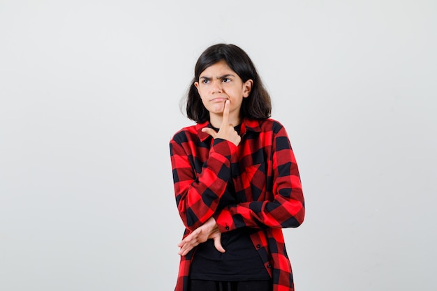 Chica adolescente sosteniendo el dedo cerca de la boca en camiseta, camisa a cuadros y mirando pensativo. vista frontal.