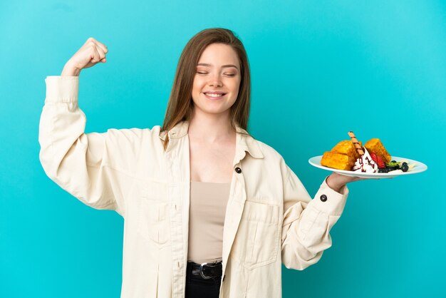 Chica adolescente sosteniendo gofres sobre fondo azul aislado haciendo un gesto fuerte