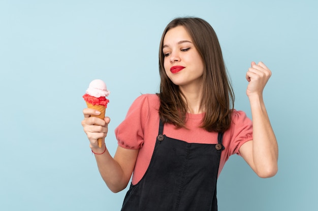 Chica adolescente sosteniendo un helado de cucurucho aislado en la pared azul celebrando una victoria