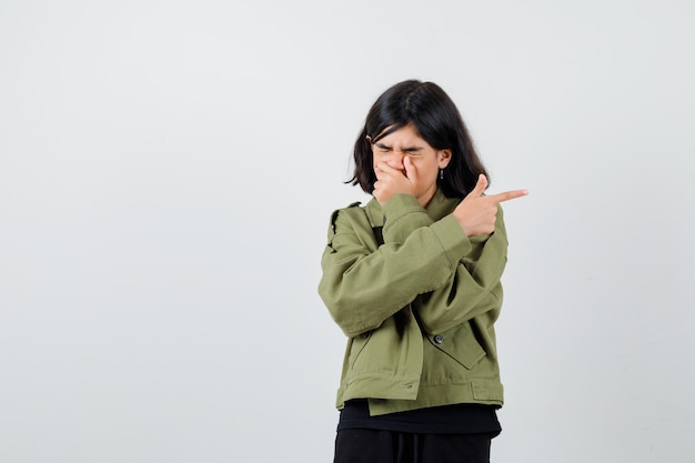 Chica adolescente sosteniendo la mano en la boca mientras apunta hacia el lado derecho en camiseta, chaqueta y mirando triste. vista frontal.