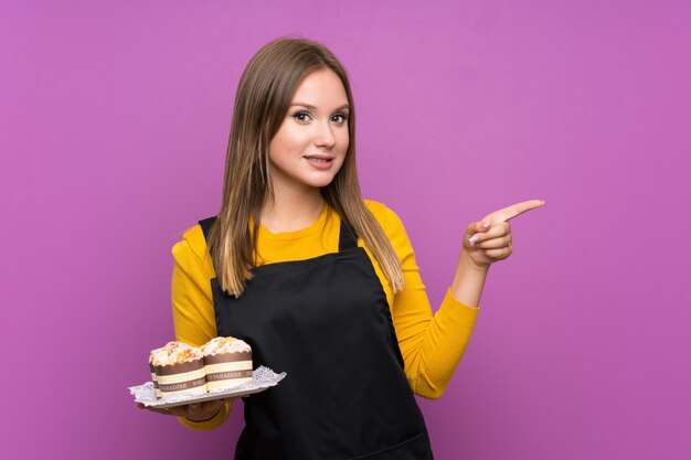 Chica adolescente sosteniendo un montón de diferentes mini pasteles sobre fondo púrpura aislado sorprendido y apuntando con el dedo hacia un lado