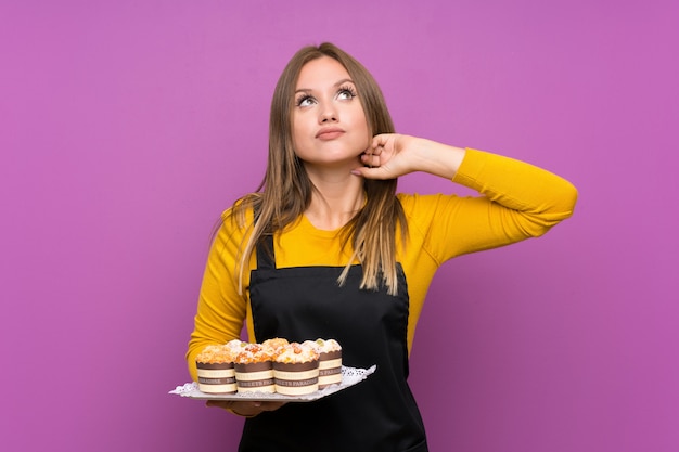 Chica adolescente sosteniendo un montón de diferentes mini tortas sobre pared púrpura aislado pensando en una idea