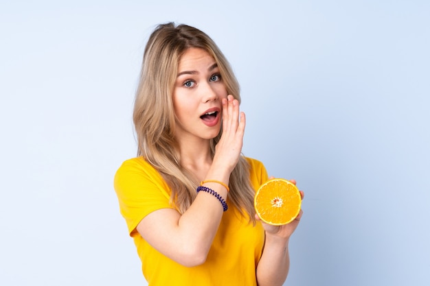 Chica adolescente sosteniendo una naranja en la pared azul susurrando algo