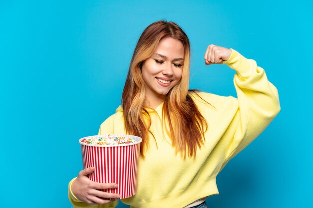 Chica adolescente sosteniendo palomitas de maíz sobre fondo azul aislado haciendo un gesto fuerte
