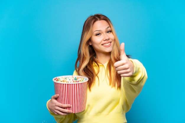 Chica adolescente sosteniendo palomitas de maíz sobre fondo azul aislado con los pulgares hacia arriba porque algo bueno ha sucedido