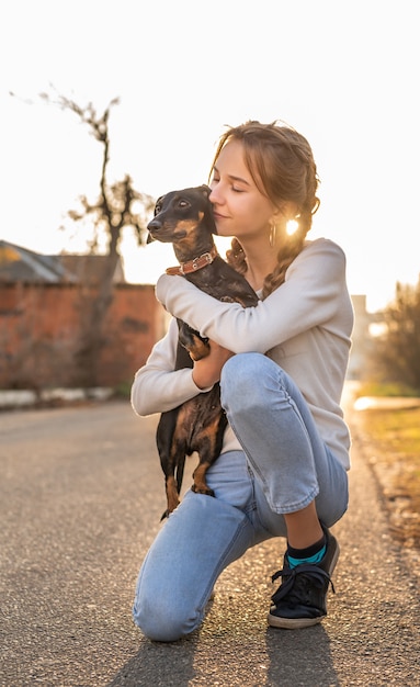 Chica adolescente sosteniendo su perro salchicha en sus brazos al aire libre en la puesta del sol