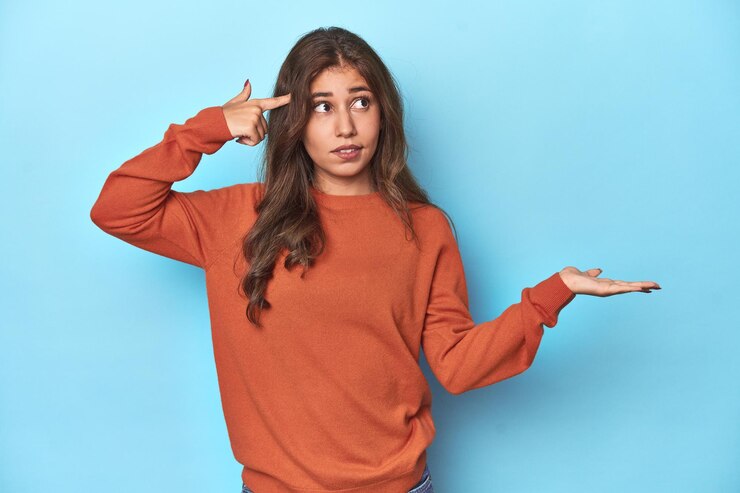 Foto chica adolescente con un suéter naranja vibrante sobre azul sosteniendo y mostrando un producto en la mano