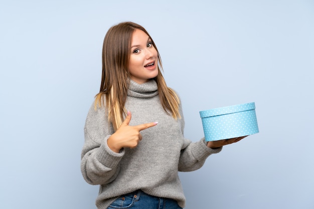 Chica adolescente con suéter sobre pared azul aislada con caja de regalo