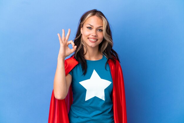 Chica adolescente superhéroe aislada sobre fondo azul mostrando signo ok con los dedos