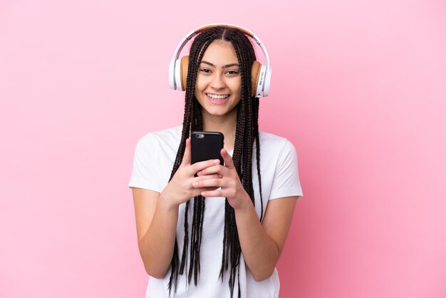 Chica adolescente con trenzas sobre fondo rosa aislado escuchando música con un móvil y mirando al frente