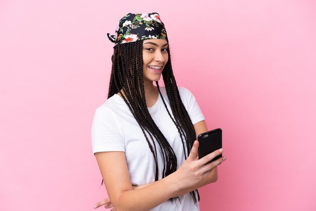 Chica adolescente con trenzas sobre fondo rosa aislado sosteniendo un teléfono móvil y con los brazos cruzados