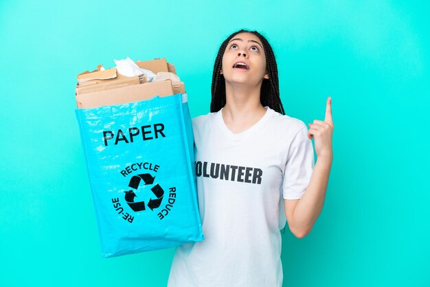 Chica adolescente con trenzas sosteniendo una bolsa para reciclar apuntando hacia arriba y sorprendida