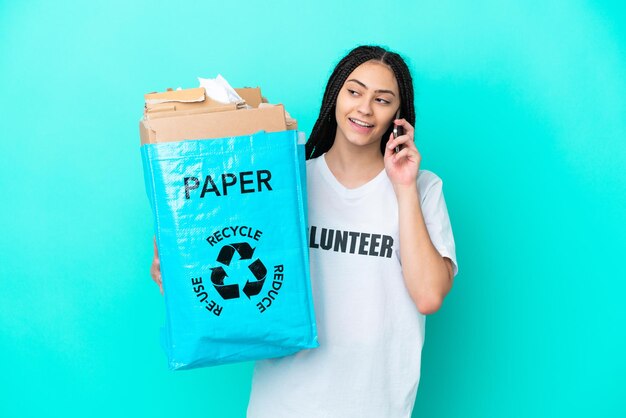 Chica adolescente con trenzas sosteniendo una bolsa para reciclar manteniendo una conversación con alguien con el teléfono móvil