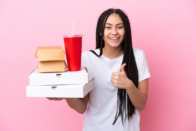 Chica adolescente con trenzas sosteniendo pizzas y hamburguesas sobre fondo rosa aislado dando un gesto de pulgar hacia arriba