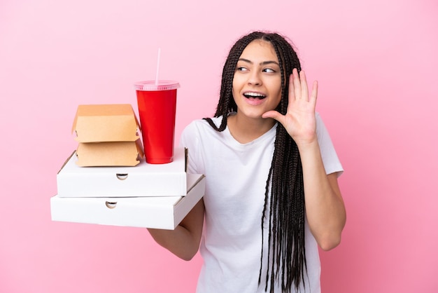 Chica adolescente con trenzas sosteniendo pizzas y hamburguesas sobre fondo rosa aislado escuchando algo poniendo la mano en la oreja