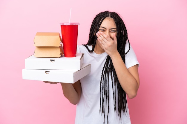Chica adolescente con trenzas sosteniendo pizzas y hamburguesas sobre fondo rosa aislado feliz y sonriente cubriendo la boca con la mano