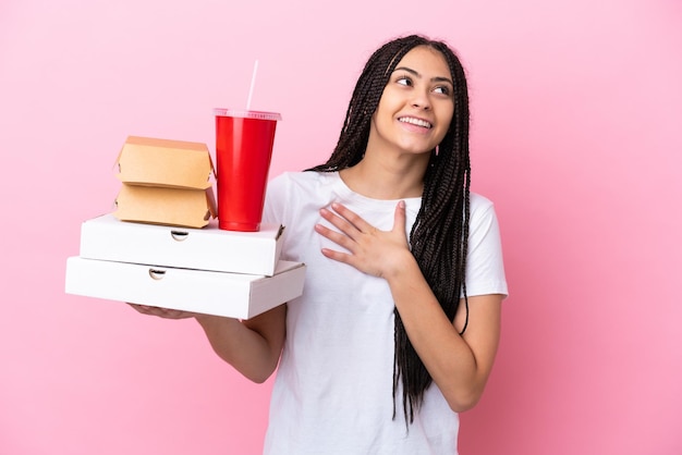 Chica adolescente con trenzas sosteniendo pizzas y hamburguesas sobre fondo rosa aislado mirando hacia arriba mientras sonríe