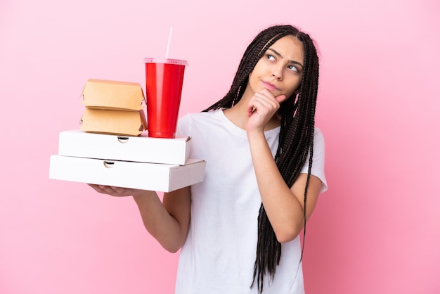 Chica adolescente con trenzas sosteniendo pizzas y hamburguesas sobre fondo rosa aislado y mirando hacia arriba