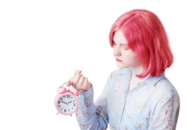 Chica adolescente triste con cabello rosado con reloj despertador aislado en blanco