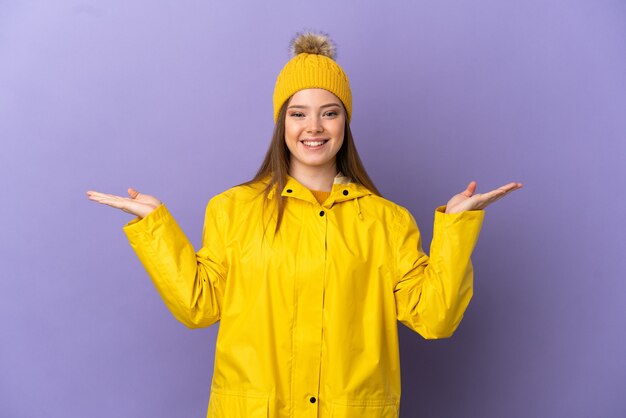 Chica adolescente vistiendo un abrigo impermeable sobre fondo púrpura aislado con expresión facial conmocionada