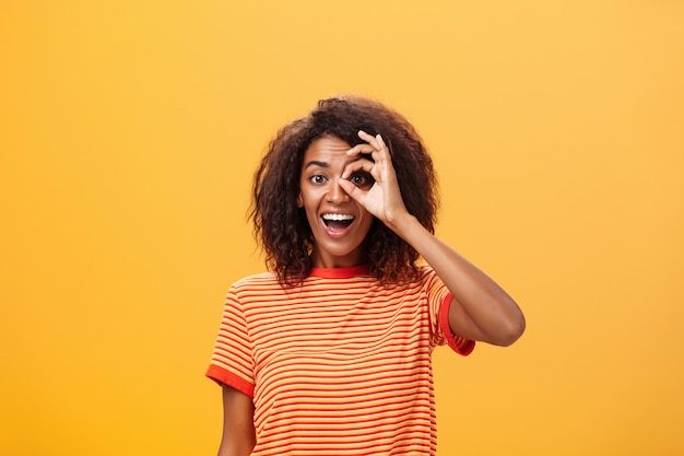 Chica adulta de piel oscura con peinado rizado en camiseta a rayas que muestra un círculo sobre el ojo o un gesto bien en la pared naranja