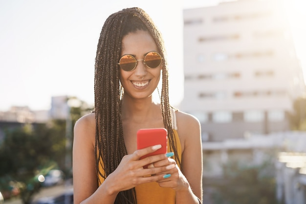 Chica africana bohemia mediante teléfono móvil al aire libre con la ciudad de fondo - Centrarse en la cara